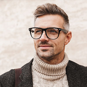 The image features a man with short hair, wearing glasses and a turtleneck sweater. He has a beard and mustache, and is looking directly at the camera.